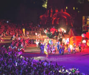 Foto de um festival noturno com muitas luzes coloridas, fogo e pessoas se apresentando