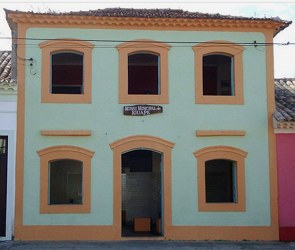 Prédio de dois andares com cinco janelas e uma porta. O prédio é branco com detalhates em amarelo em volta das janelas.