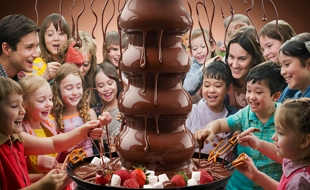 Imagem de crian&ccedil;as e adultos comendo chocolate numa torre de chocolate derretido.