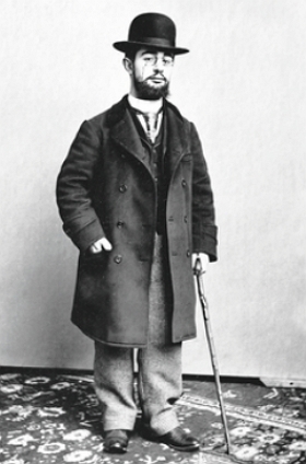 Foto de corpo inteiro de Toulouse-Lautrec, homem branco de barba e bigode, usando roupa preta e chapéu preto