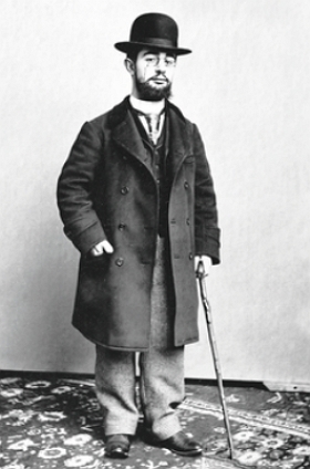Foto de corpo inteiro de Toulouse-Lautrec, homem branco de barba e bigode, usando roupa preta e chap&eacute;u preto