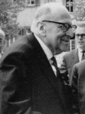 Foto de um homem idoso, de perfil, com óculos preto, usando terno e gravata