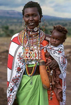 Foto de uma mulher massai com roupas coloridas, segurando um filho no colo.